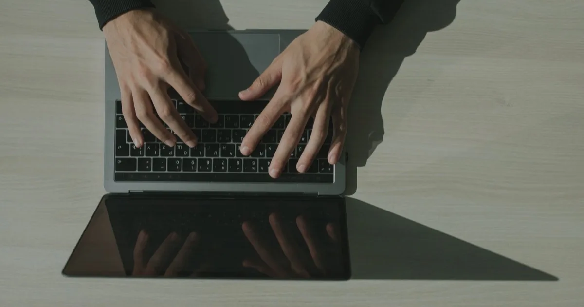 Hands typing on a laptop viewed from above, reflected in the dark screen.