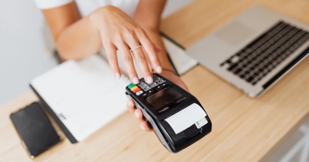 A person entering a number on a handheld payment terminal beside a laptop.