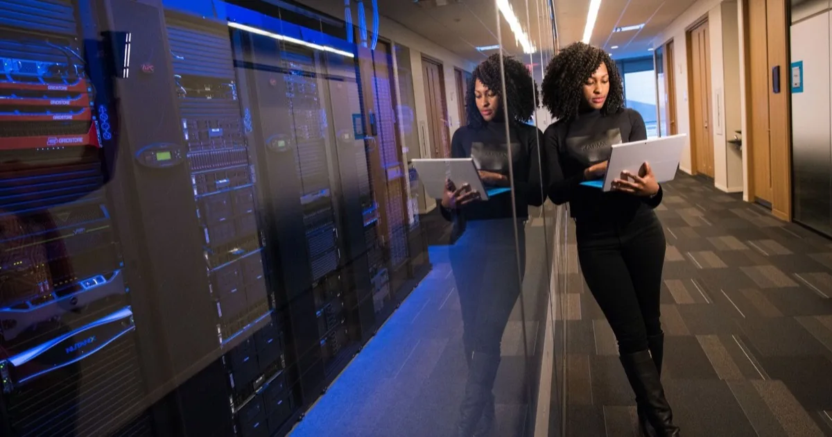 An engineer carrying a laptop beside glass-fronted server racks.