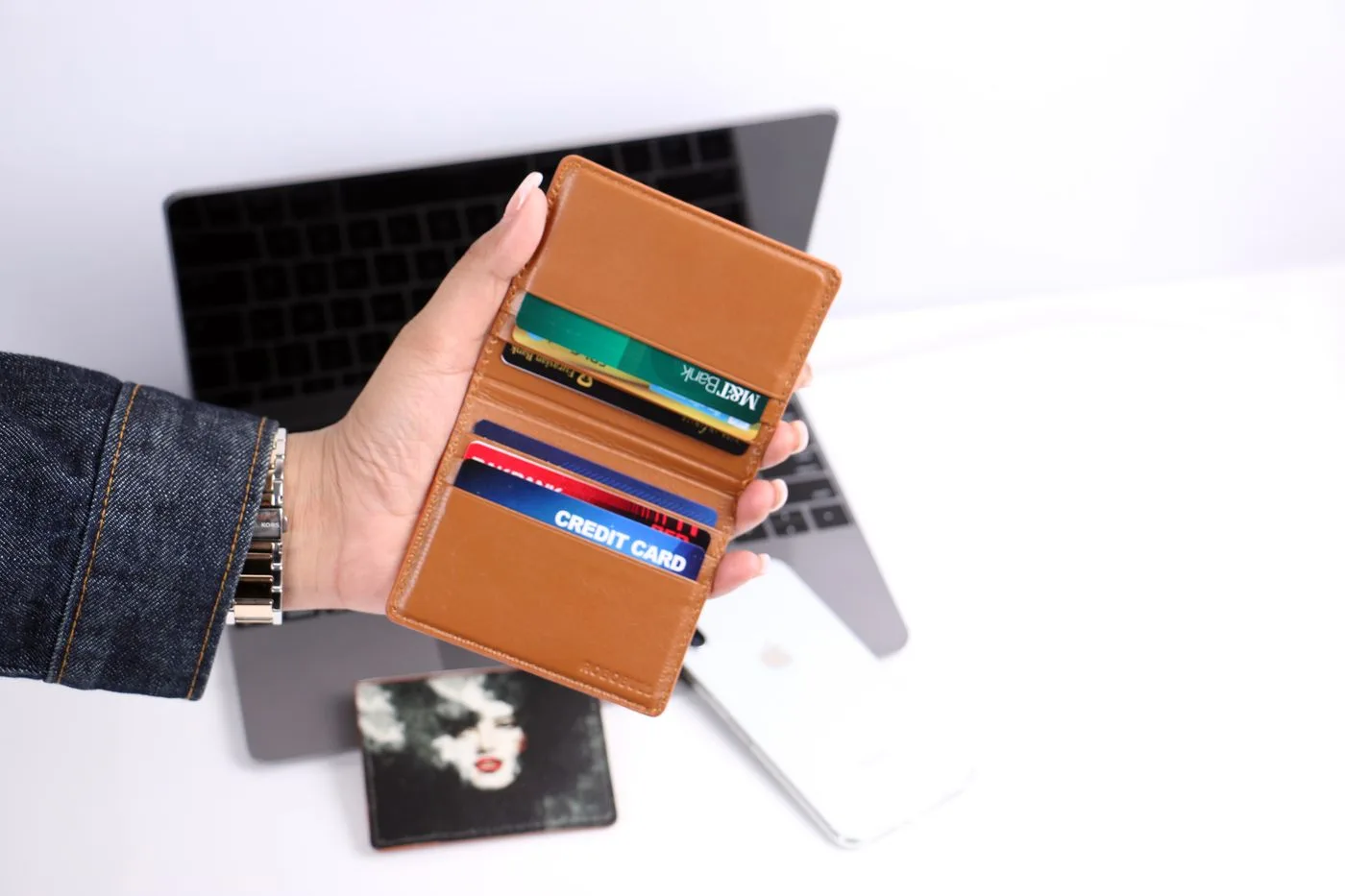 A hand holds a wallet with several bank cards in front of a laptop.