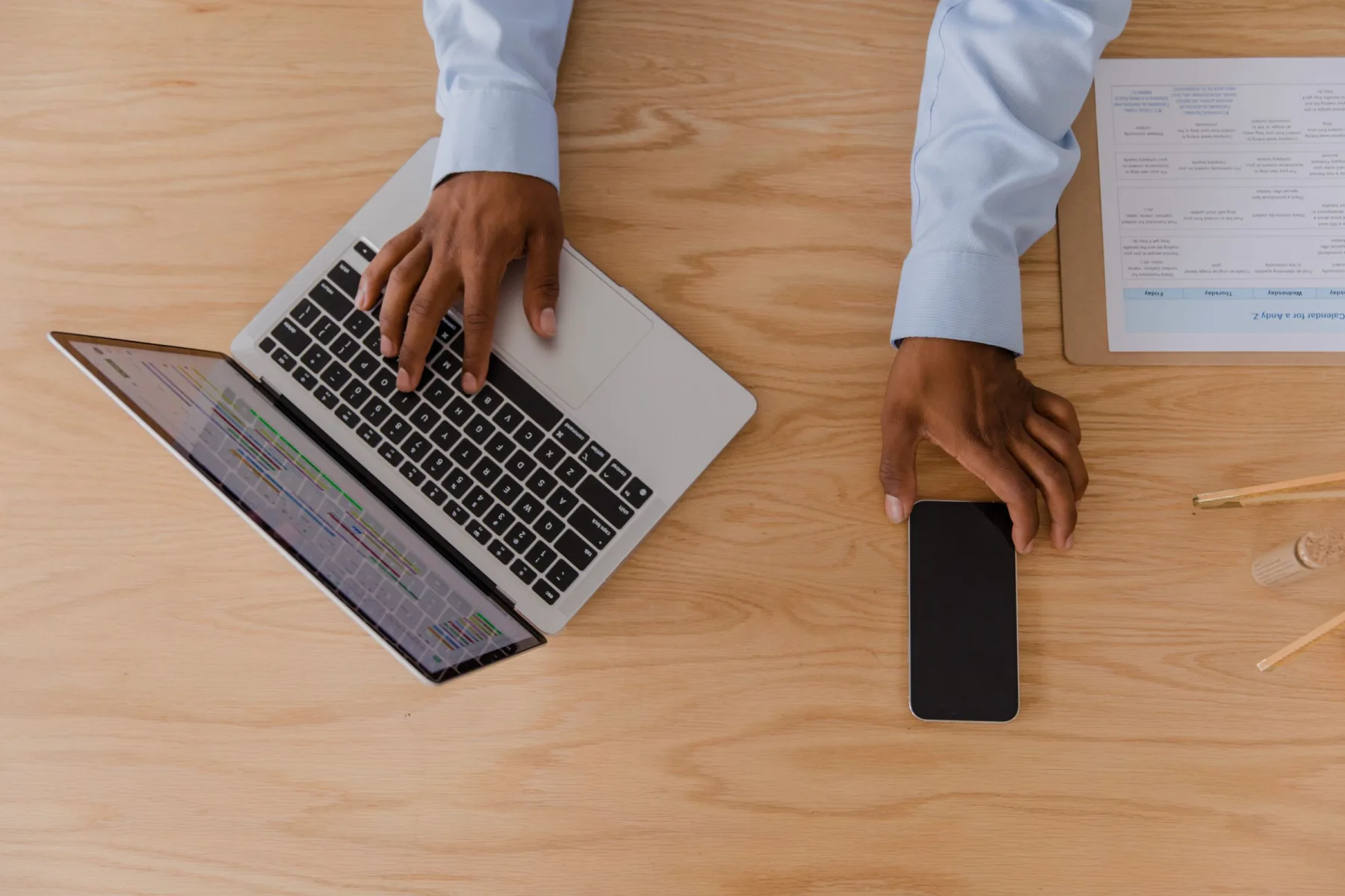 Overhead view of a person using a laptop and reaching for a phone on a wooden desk.